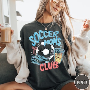 May include: Dark gray sweatshirt with the words "Soccer Moms Club" in blue and pink. The design includes a soccer ball, cleats, and a drink with a straw. The sweatshirt is a relaxed fit.