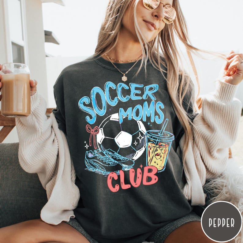 May include: Dark gray sweatshirt with the words "Soccer Moms Club" in blue and pink. The design includes a soccer ball, cleats, and a drink with a straw. The sweatshirt is a relaxed fit.