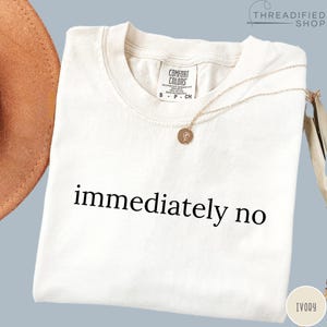 May include: Ivory-colored t-shirt with the phrase "immediately no" printed in black. A gold necklace with a pendant rests on the shirt. The shirt has a Comfort Colors tag.