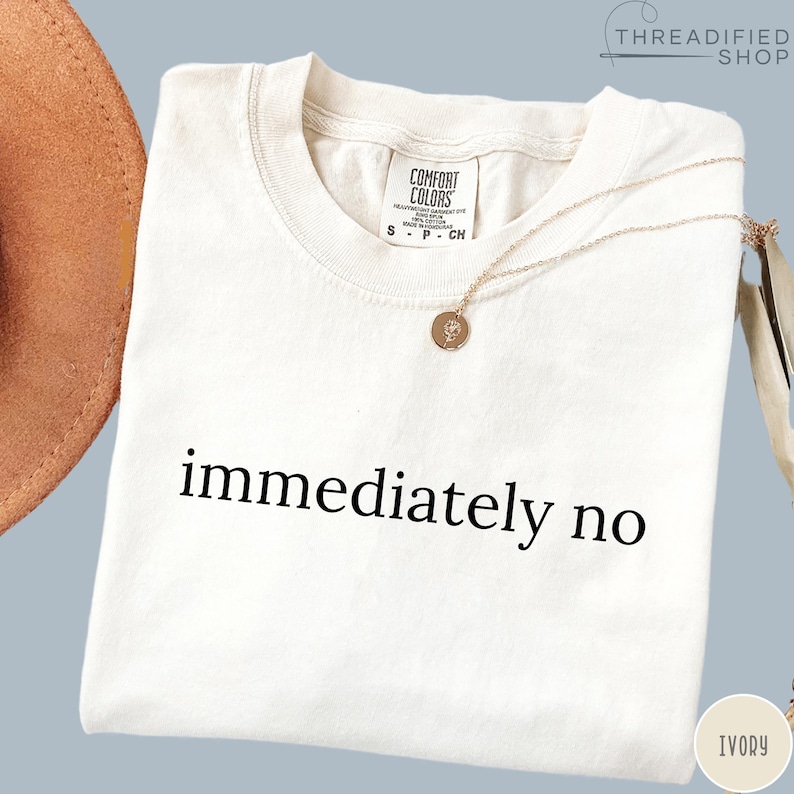 May include: Ivory-colored t-shirt with the phrase "immediately no" printed in black. A gold necklace with a pendant rests on the shirt. The shirt has a Comfort Colors tag.