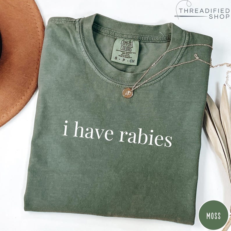 May include: A moss green t-shirt with the phrase "i have rabies" printed in white. The shirt has a crew neck and short sleeves. A gold necklace with a pendant is draped across the shirt.