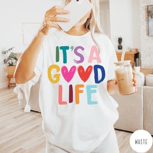 K&ouml;nnte beinhalten: Ein wei&szlig;es Kurzarm-T-Shirt mit dem bunten Schriftzug IT'S A GOOD LIFE auf der Vorderseite. Die Buchstaben sind in verschiedenen leuchtenden Farben wie Gr&uuml;n, Lila, Pink, Gelb, Rot, Blau und Orange gehalten und werden durch zwei Herzformen erg&auml;nzt.