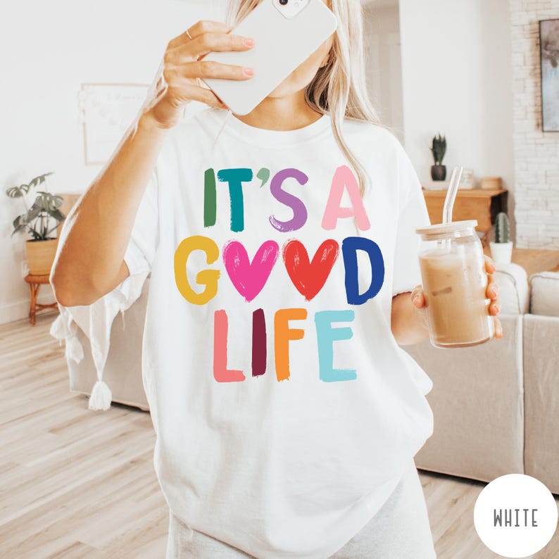 K&ouml;nnte beinhalten: Ein wei&szlig;es Kurzarm-T-Shirt mit dem bunten Schriftzug IT'S A GOOD LIFE auf der Vorderseite. Die Buchstaben sind in verschiedenen leuchtenden Farben wie Gr&uuml;n, Lila, Pink, Gelb, Rot, Blau und Orange gehalten und werden durch zwei Herzformen erg&auml;nzt.