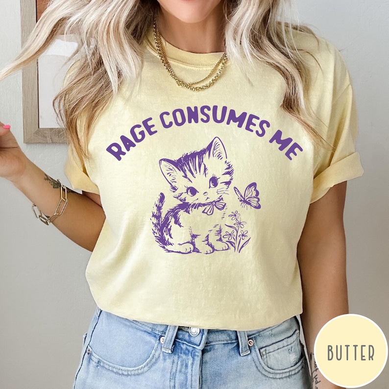 Puede incluir: Una camiseta amarilla con el texto "Rage Consumes Me" impreso en morado. Un gatito de dibujos animados morado con un lazo est&aacute; en el centro de la camiseta, con una mariposa y flores.