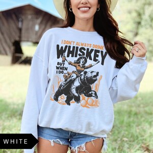 May include: White sweatshirt with a graphic print of a woman riding a bear and holding a bottle of whiskey. The text on the shirt reads "I don't always drink whiskey but when I do..."