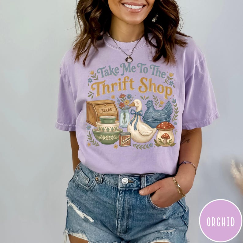 Op de afbeelding: Een lila T-shirt met de tekst "Take Me To The Thrift Shop" en een vintage illustratie. De afbeelding toont een brooddoos, kommen, een eend, een blauwe kip en een paddenstoel.
