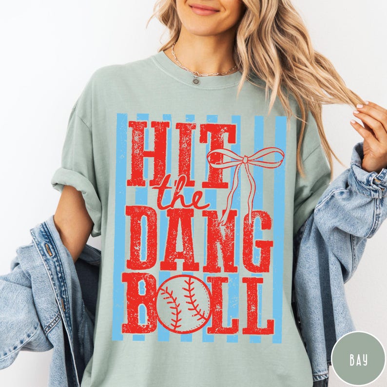Pu&ograve; includere: T-shirt verde chiaro con la scritta "HIT the DANG BALL" in rosso, insieme a una grafica di baseball. Il design &egrave; ambientato su uno sfondo di strisce verticali azzurre. &Egrave; incluso anche un fiocco rosso.