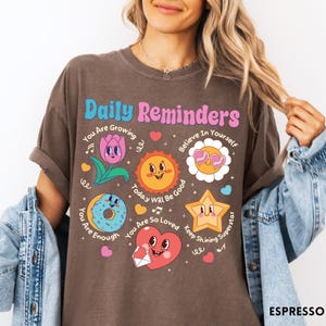 May include: Espresso-colored t-shirt with the text "Daily Reminders" and various cartoon graphics. The graphics include a flower, sun, donut, star, and heart, each with positive affirmations. The shirt is a relaxed fit.