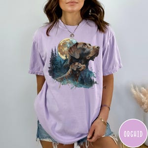 May include: A light purple t-shirt featuring a watercolor illustration of two brown dogs with a full moon and forest background. The word "ORCHID" is printed on a pink circle in the lower right corner.