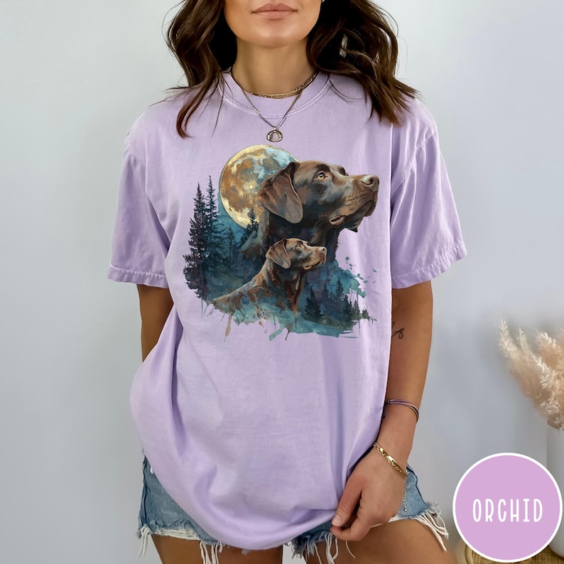 May include: A light purple t-shirt featuring a watercolor illustration of two brown dogs with a full moon and forest background. The word "ORCHID" is printed on a pink circle in the lower right corner.