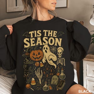 Puede incluir: Sudadera negra con un dise&ntilde;o de Halloween. El dise&ntilde;o incluye una calabaza, un fantasma, una calavera, un sombrero de bruja y el texto "TIS THE SEASON" en un estilo vintage. Tambi&eacute;n hay una casa embrujada, botellas de pociones y una l&aacute;pida.