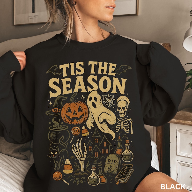Puede incluir: Sudadera negra con un dise&ntilde;o de Halloween. El dise&ntilde;o incluye una calabaza, un fantasma, una calavera, un sombrero de bruja y el texto "TIS THE SEASON" en un estilo vintage. Tambi&eacute;n hay una casa embrujada, botellas de pociones y una l&aacute;pida.