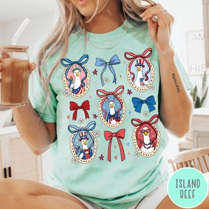 May include: A light green t-shirt featuring a repeating pattern of patriotic goose illustrations within decorative frames. Each goose is adorned with American flag-themed accessories and surrounded by bows and floral accents. The shirt has a relaxed fit.
