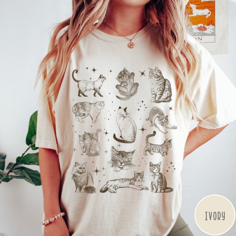 K&ouml;nnte beinhalten: Ein cremefarbenes T-Shirt mit einem grafischen Druck verschiedener Katzen in verschiedenen Posen. Die Katzen sind alle in Schwarzwei&szlig; und von Sternen umgeben. Das Shirt ist mit "IVORY" gekennzeichnet.