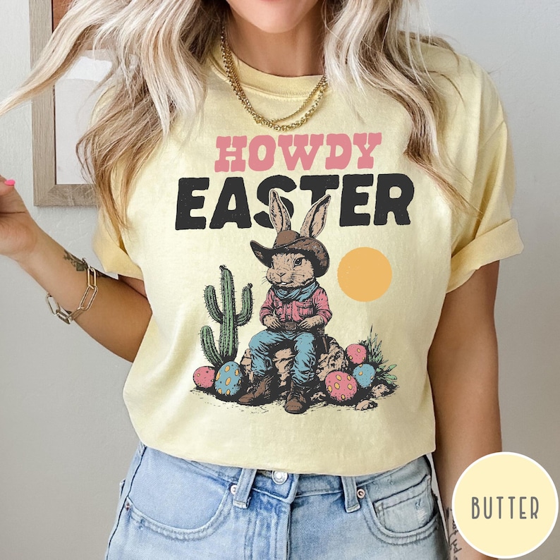 Puede incluir: Camiseta amarilla clara con las palabras "HOWDY EASTER" en rosa y negro. El gr&aacute;fico presenta un conejo con sombrero de vaquero, sentado en una roca con huevos de Pascua y un cactus. Un c&iacute;rculo amarillo representa el sol.
