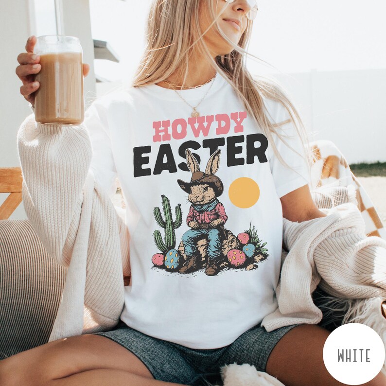 Puede incluir: Camiseta blanca con la frase "HOWDY EASTER" en rosa y negro. El dise&ntilde;o presenta un conejito con sombrero de vaquero sentado en una roca con huevos de Pascua y un cactus. Un c&iacute;rculo amarillo est&aacute; a la derecha del conejo.