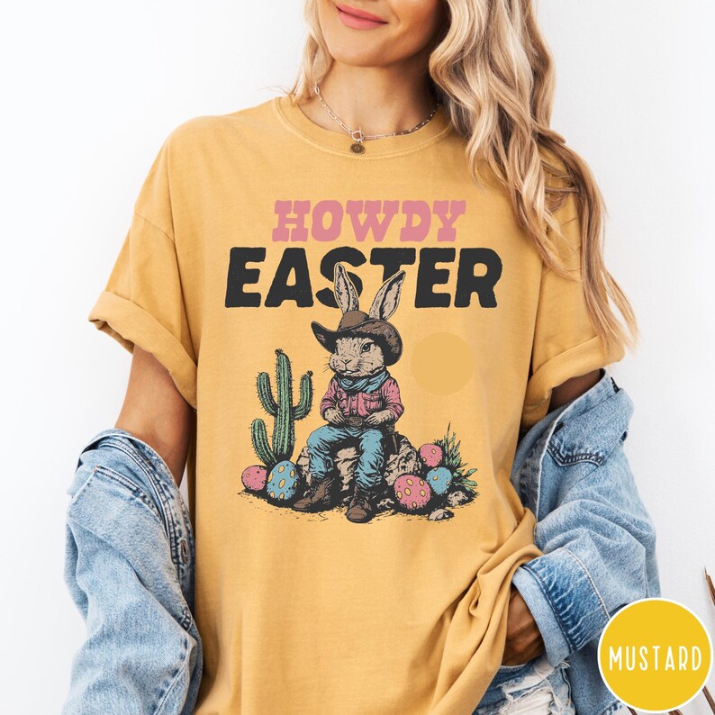 Puede incluir: Camiseta amarilla mostaza con las palabras "HOWDY EASTER" en rosa y negro. El gr&aacute;fico presenta un conejito con sombrero de vaquero y camisa a cuadros, sentado en una roca con huevos de Pascua y un cactus. La camiseta es de estilo casual y unisex.