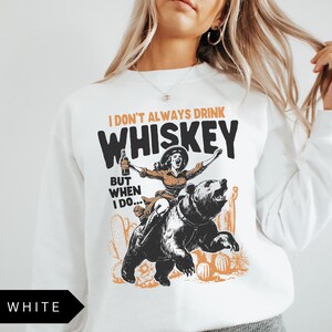 May include: White sweatshirt with a graphic of a woman riding a bear and holding a bottle of whiskey. The text on the shirt reads "I don't always drink whiskey, but when I do..."