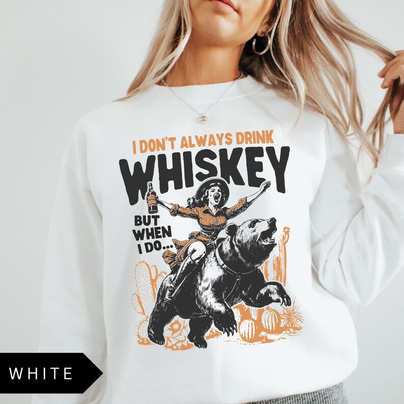 May include: White sweatshirt with a graphic of a woman riding a bear and holding a bottle of whiskey. The text on the shirt reads "I don't always drink whiskey, but when I do..."
