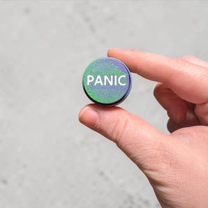 May include: A circular button with the word "PANIC" in white text, set against a green and purple background. The button is held between two fingers against a light grey backdrop.