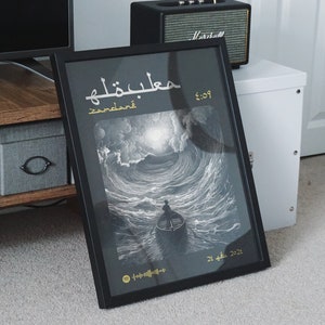 May include: A black framed print with a black and white illustration of a person in a boat on a stormy sea. The print has gold text that reads "Maelstrom" and "5:09" in Arabic and English. The print also has a Spotify code and the date "21. 4. 2021".