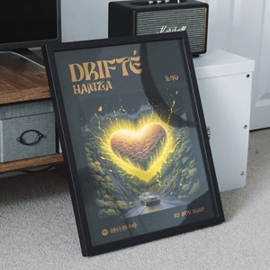 May include: A framed art print featuring the text "DRIFTÉ HANZA" in gold lettering. The artwork depicts a glowing heart shape over a road winding through a forest, with a car driving on it. The date "23 NOV 2023" is also visible.