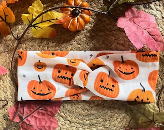 May include: White headband with a twisted knot design. The headband is covered in a repeating pattern of orange jack-o'-lantern faces on a white background.