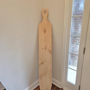 Peut inclure: Une grande planche en bois clair non fini avec un motif de grain de bois naturel. La planche présente une poignée en haut avec une découpe en forme de losange et une base pointue avec un design cranté décoratif sur les deux côtés.