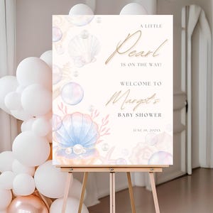 May include: A white sign with a watercolor design of seashells, pearls, and bubbles. The text reads "A Little Pearl is on the Way!" and "Welcome to Marget's Baby Shower June 19, 202XX".