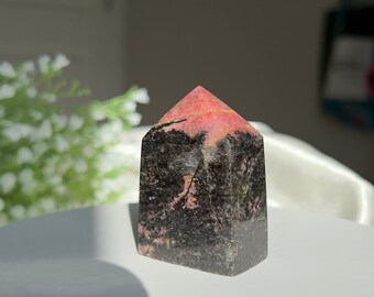 Puede incluir: Una punta de cristal pulida de color rosa y negro, también conocida como cristal de rodonita, con una parte superior puntiaguda y una base plana.
