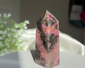 Puede incluir: Una punta de cristal rosa y negra con una parte superior puntiaguda y una base hexagonal. El cristal tiene una textura natural y rugosa.