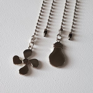 Puede incluir: Tres cadenas de tirador de ventilador de techo plateadas con diferentes extremos decorativos. Un extremo tiene forma de bombilla, otro tiene forma de aspa de ventilador y el último es un simple bucle.