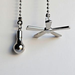 May include: Two silver-toned ceiling fan and light pull chains hanging against a white background. One pull chain has a light bulb charm, and the other has a fan blade charm. Both charms are suspended from a beaded chain.