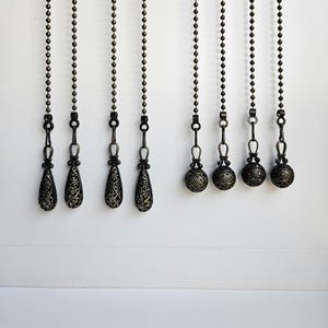 May include: Decorative bronze-colored ceiling fan pull chains. The chains have ornate, filigree-style teardrop and ball-shaped pulls. The pulls hang from a beaded chain against a white background. The design adds a touch of elegance to any room.
