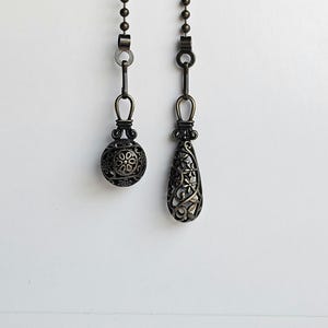 May include: Two decorative bronze-colored ceiling fan pull chains. One is a sphere with a floral design, and the other is a teardrop shape with a floral and star pattern. Both are suspended from a beaded chain.