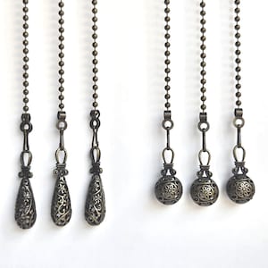 Puede incluir: Seis cadenas decorativas de bronce para ventiladores de techo o lámparas. Las cadenas tienen colgantes ornamentados de estilo filigrana en forma de lágrima y esférica. Cada cadena cuelga sobre un fondo blanco.
