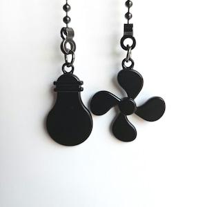 May include: Two black ceiling fan and light pull chain ornaments. One is shaped like a lightbulb, and the other is a three-blade fan. Both are suspended from a beaded chain against a white background.