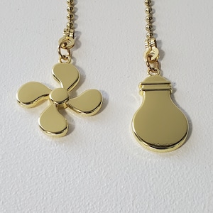 Puede incluir: Dos cadenas de tirador de ventilador doradas, una con un diseño de aspa de ventilador y la otra con un diseño de bombilla.