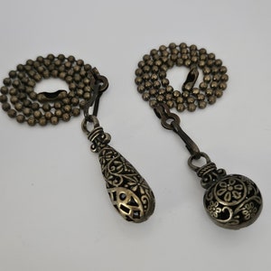 Puede incluir: Dos cadenas de tirador de ventilador de bronce decorativas con diseños ornamentados. Una cadena tiene un tirador en forma de lágrima con un patrón floral, la otra tiene un tirador redondo con un patrón floral.