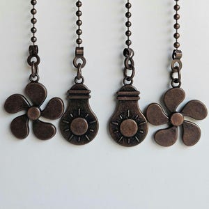 Puede incluir: Cuatro cadenas de tirador de ventilador de techo de color bronce con colgantes decorativos. Dos colgantes tienen forma de flor y dos de bombilla. Cada colgante cuelga de una cadena de cuentas.