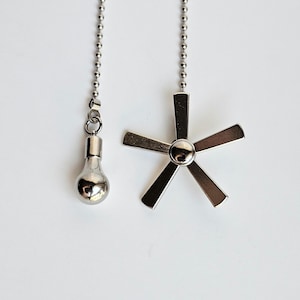 May include: Two decorative ceiling fan pull chains. One chain has a light bulb shaped pull, and the other has a fan blade shaped pull. Both pulls are silver and hang from a silver ball chain against a white background.