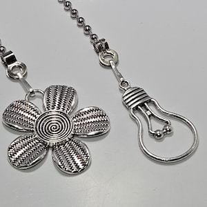 Puede incluir: Una cadena de plata con un dije de flor y bombilla. La cadena está hecha de pequeñas cuentas de metal.