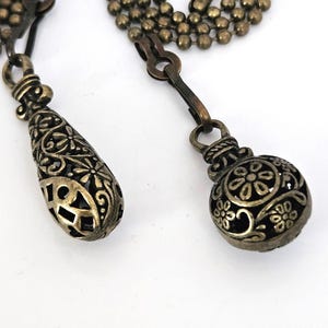 May include: Close-up of two antique bronze-toned pendants. One is a teardrop shape with openwork filigree, the other is a round ball with floral cutouts. Both are attached to a ball chain and a clasp, set against a white background.