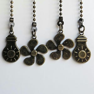 Puede incluir: Cuatro adornos de cadena de tracción de ventilador de techo y luz de bronce antiguo. Dos tienen forma de bombillas con diseños de sol, y dos tienen forma de flor. Cada adorno cuelga de una cadena de cuentas.