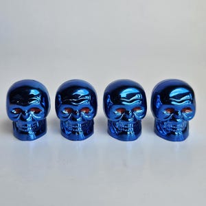 May include: Four metallic blue skull figurines with red eye sockets are arranged in a row. The skulls have a glossy finish and are set against a plain white background. The skulls are a decorative item.