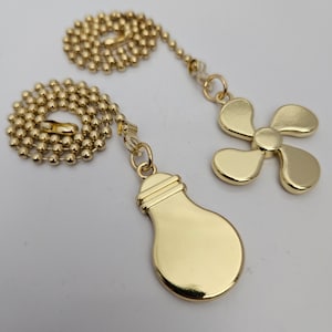 Puede incluir: Dos cadenas de tirador de luz de color dorado con extremos decorativos. Un extremo tiene forma de bombilla y el otro tiene forma de ventilador.