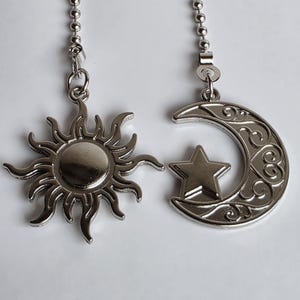 Puede incluir: Juego de tiradores de ventilador de techo de sol y luna plateados. El sol tiene rayos radiantes y un centro circular. La luna presenta una estrella y remolinos decorativos. Ambos están unidos a cadenas de cuentas.