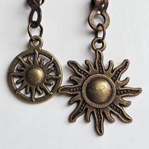 May include: Two antique bronze-colored earrings. One earring features a circular sunburst design, while the other showcases a detailed sun pendant with radiating rays. Both earrings hang from a chain with a small ball at the top.