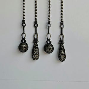 Puede incluir: Cuatro cadenas de tirador de ventilador de color bronce antiguo con colgantes decorativos de filigrana. Los colgantes tienen diferentes formas, incluyendo redondas y de lágrima, y están suspendidos de cadenas de cuentas sobre un fondo blanco.