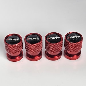 May include: Four red metal tire valve stem caps with a textured design. Each cap has a black top with the word "SPORT" in white. The caps are designed for automotive use.